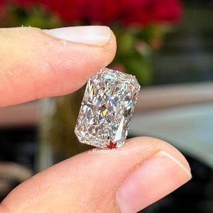 May include: A rectangular-cut diamond held between fingers. The diamond sparkles with internal reflections, showcasing its facets. The background is blurred, with red flowers visible. The text "Minima Jewelry" is visible on the diamond.