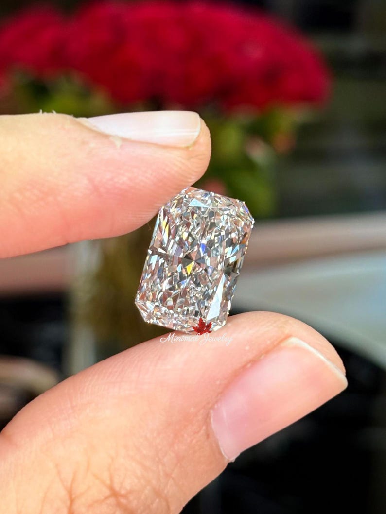 May include: A rectangular-cut diamond held between fingers. The diamond sparkles with internal reflections, showcasing its facets. The background is blurred, with red flowers visible. The text "Minima Jewelry" is visible on the diamond.