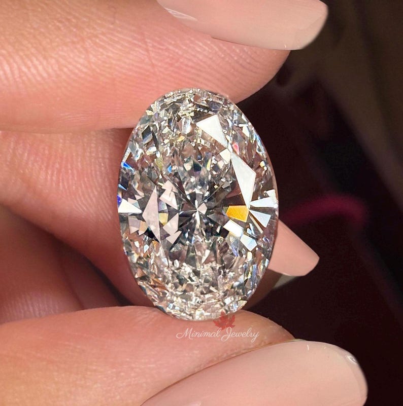 May include: A large, oval-cut diamond with numerous facets, held between fingers. The diamond sparkles with internal reflections, showcasing its clarity and brilliance. The text "Minimal Jewelry" is visible at the bottom.