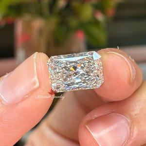 May include: A rectangular, clear diamond with multiple facets, held between fingers. The diamond reflects light, creating a sparkling effect. The background is blurred, with red flowers visible. The text "Minimal Jewelry" is in the lower left corner.