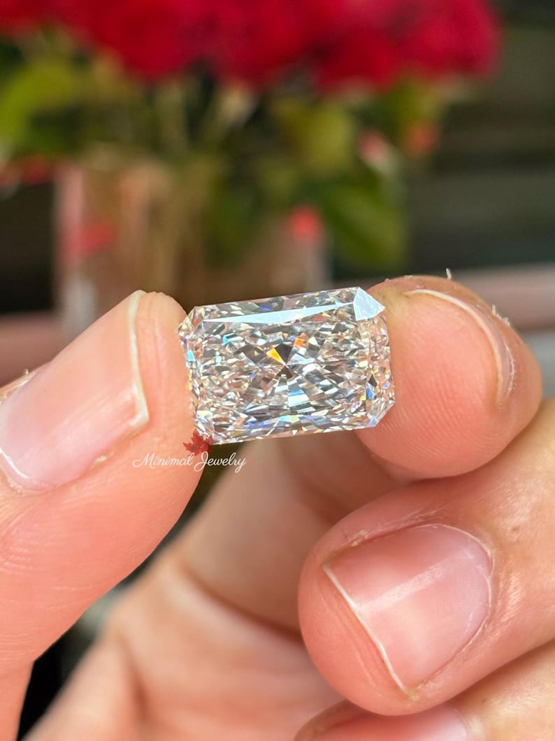 May include: A rectangular, clear diamond with multiple facets, held between fingers. The diamond reflects light, creating a sparkling effect. The background is blurred, with red flowers visible. The text "Minimal Jewelry" is in the lower left corner.
