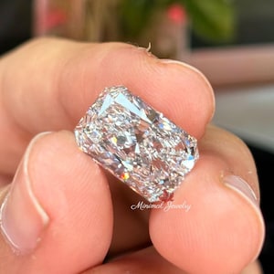 May include: A radiant-cut diamond held between fingers. The diamond is rectangular with many facets, reflecting light. The image is close-up, highlighting the clarity and brilliance of the gemstone. The text "Minimal Jewelry" is visible.