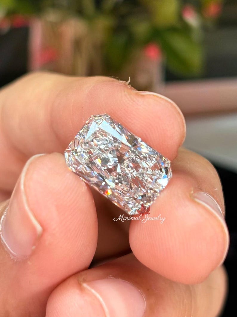 May include: A radiant-cut diamond held between fingers. The diamond is rectangular with many facets, reflecting light. The image is close-up, highlighting the clarity and brilliance of the gemstone. The text "Minimal Jewelry" is visible.
