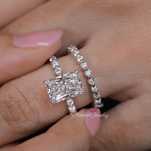 May include: A radiant-cut diamond engagement ring and a diamond band. The engagement ring has a large, rectangular diamond on a slender band. The band is decorated with smaller diamonds. The rings are shown on a hand with pink nail polish.
