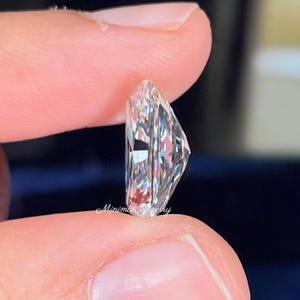 May include: A clear, radiant-cut diamond held between fingers. The gemstone has a rectangular shape with multiple facets, reflecting light. The image is a close-up, highlighting the clarity and brilliance of the diamond. The text "Minimal Jewelry" is visible.