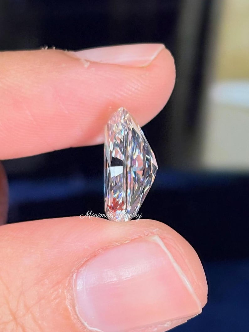 May include: A clear, radiant-cut diamond held between fingers. The gemstone has a rectangular shape with multiple facets, reflecting light. The image is a close-up, highlighting the clarity and brilliance of the diamond. The text "Minimal Jewelry" is visible.