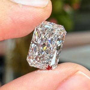 May include: A large, rectangular-cut diamond with brilliant facets is held between fingers. The diamond sparkles with internal reflections, showcasing its clarity and cut. The text "Minimal Jewelry" is visible on the bottom of the diamond.