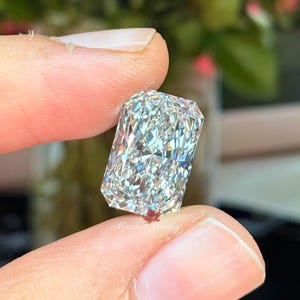 May include: A radiant-cut diamond held between fingers. The rectangular gemstone sparkles with numerous facets, reflecting light. The diamond is clear and colorless, showcasing its brilliance. The background is blurred, with hints of red and green.