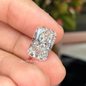 May include: A radiant-cut diamond with a rectangular shape, held between fingers. The diamond sparkles with internal reflections, showcasing its facets. The image is well-lit, highlighting the clarity and brilliance of the gemstone. The text "Minimal Jewelry" is visible.