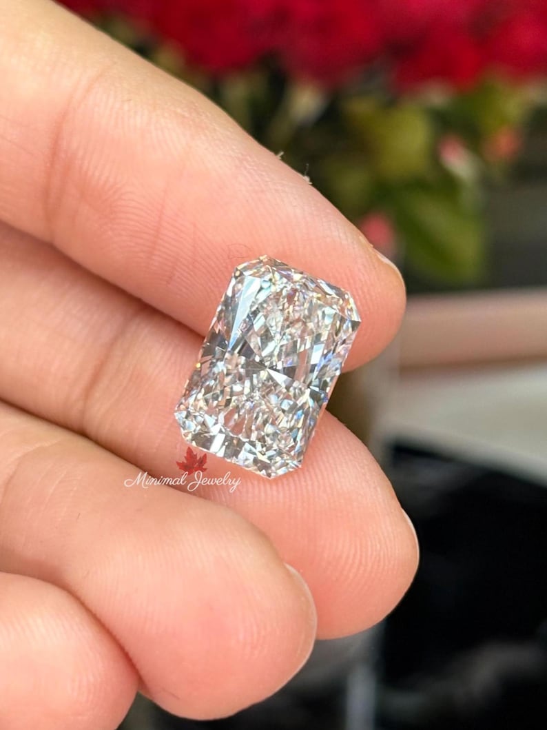 May include: A radiant-cut diamond with a rectangular shape, held between fingers. The diamond sparkles with internal reflections, showcasing its facets. The image is well-lit, highlighting the clarity and brilliance of the gemstone. The text "Minimal Jewelry" is visible.