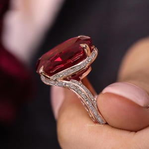 Könnte beinhalten: Ein roségoldener Ring mit einem großen, oval geschliffenen, tiefroten Edelstein. Der Edelstein ist von einem Kranz aus kleineren, rechteckigen, klaren Steinen umgeben. Auch das Ringband weist die kleineren Steine auf.