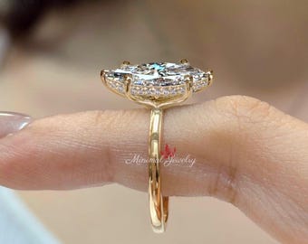 May include: A gold ring featuring a large marquise-cut center stone set in a basket with a hidden halo of small pavé diamonds. The band is smooth and polished gold. The text Minimal Jewelry is visible in red script on the side of the ring setting.