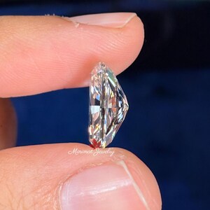 May include: A clear, elongated diamond with multiple facets is held between two fingers. The diamond is cut in a unique shape, reflecting light and creating a sparkling effect. The text "Minimal Jewelry" is visible.