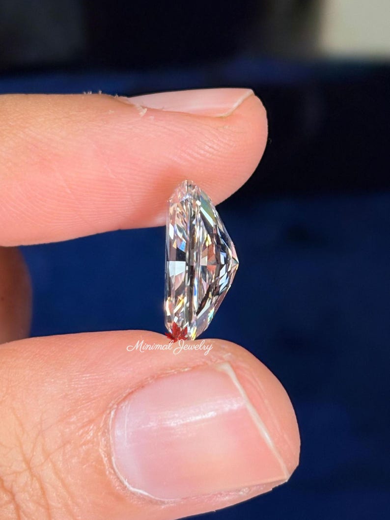 May include: A clear, elongated diamond with multiple facets is held between two fingers. The diamond is cut in a unique shape, reflecting light and creating a sparkling effect. The text "Minimal Jewelry" is visible.
