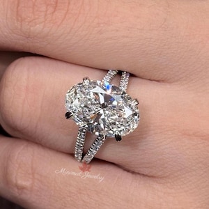 Op de afbeelding: Een zilverkleurige ring met een grote, ovaal geslepen heldere steen in het midden, vastgehouden door vier pootjes. De ringband is gespleten in twee dunne, parallelle banen, beide bezet met kleine, ronde, heldere steentjes. De tekst Minimal Jewelry is zichtbaar.