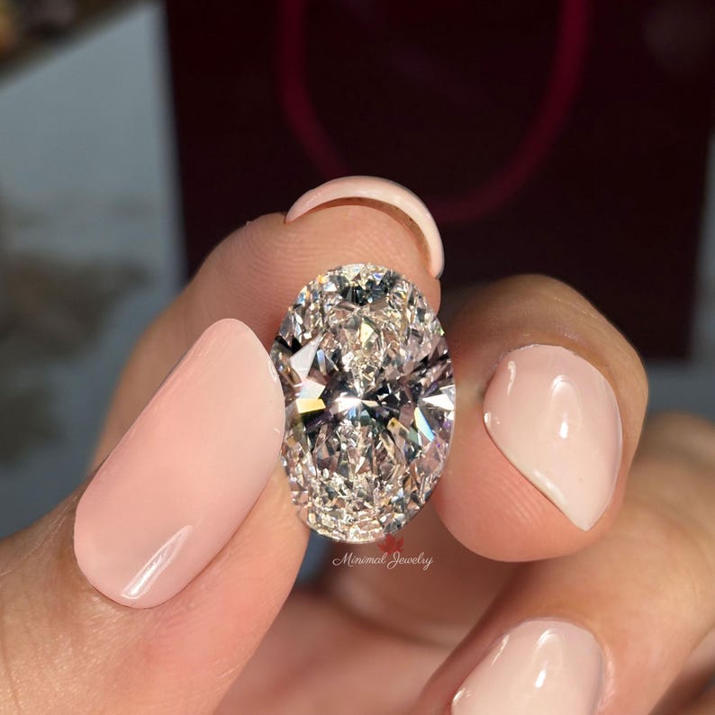 May include: An oval-cut diamond held between fingers with a light pink manicure. The diamond is clear and reflects light, showcasing its facets. The words "Minimal Jewelry" are visible on the diamond.