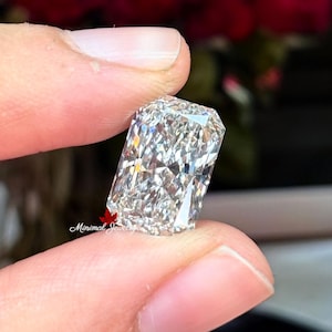 May include: A rectangular-cut diamond with a brilliant, multifaceted surface. The gem is held between fingers, showcasing its clarity and sparkle. The background features blurred red and dark tones, highlighting the diamond's brilliance. The Minimal Jewels logo is visible.