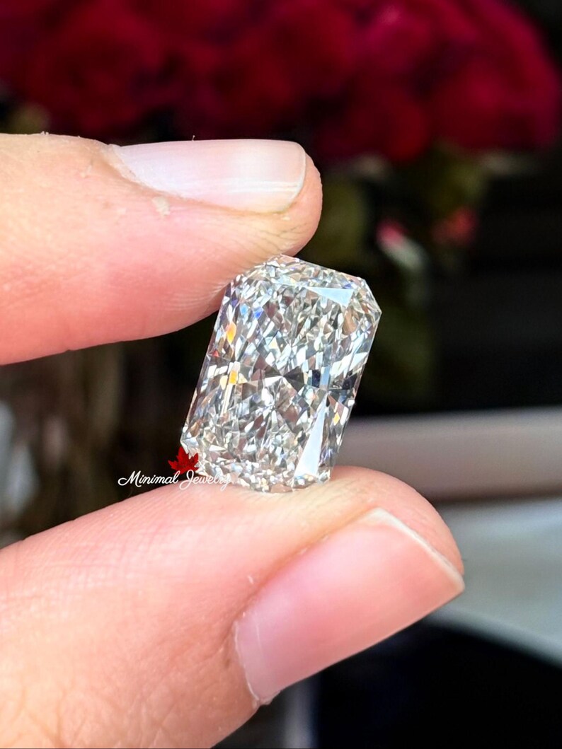 May include: A rectangular-cut diamond with a brilliant, multifaceted surface. The gem is held between fingers, showcasing its clarity and sparkle. The background features blurred red and dark tones, highlighting the diamond's brilliance. The Minimal Jewels logo is visible.