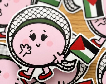 May include: A selection of stickers depicting a cartoon character with a pink face, black eyes, and red shoes. The character is wearing a white head covering and holding a Palestinian flag. The stickers are on a wooden surface.