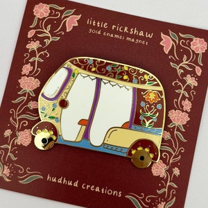 Puede incluir: Imán de esmalte dorado de un rickshaw colorido y decorado con el texto "little rickshaw gold enamel magnet" y "hudhud creations" sobre un fondo rojo con diseños florales.