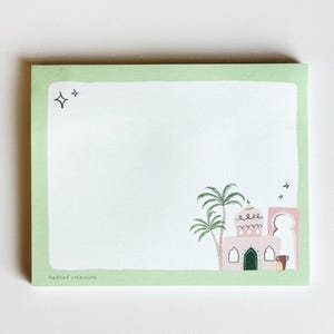 Peut inclure: Un bloc-notes carré avec une bordure vert clair et un intérieur blanc. Le bloc-notes présente une illustration fantaisiste d'un bâtiment rose avec des palmiers et une porte verte. Deux étoiles sont dans le coin supérieur gauche. Le texte "badhad creations" est en bas.