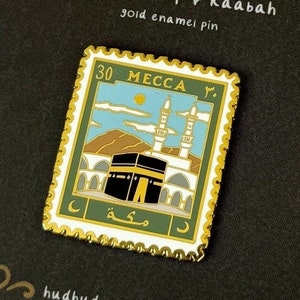 Pode incluir: Pin de esmalte dourado em forma de selo postal com fundo verde e borda branca. O selo apresenta uma representação da Caaba em Meca, Arábia Saudita, com o texto árabe "Meca" e "30" impresso no selo. O pin está sobre um fundo preto com uma borda decorativa dourada.