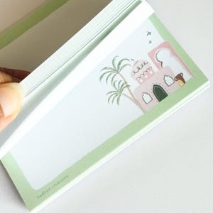 May include: A notepad with a light green border and a decorative illustration of a pink building, palm trees, and a small animal. The notepad is open, revealing blank pages. The text "hudhud creations" is printed at the bottom.