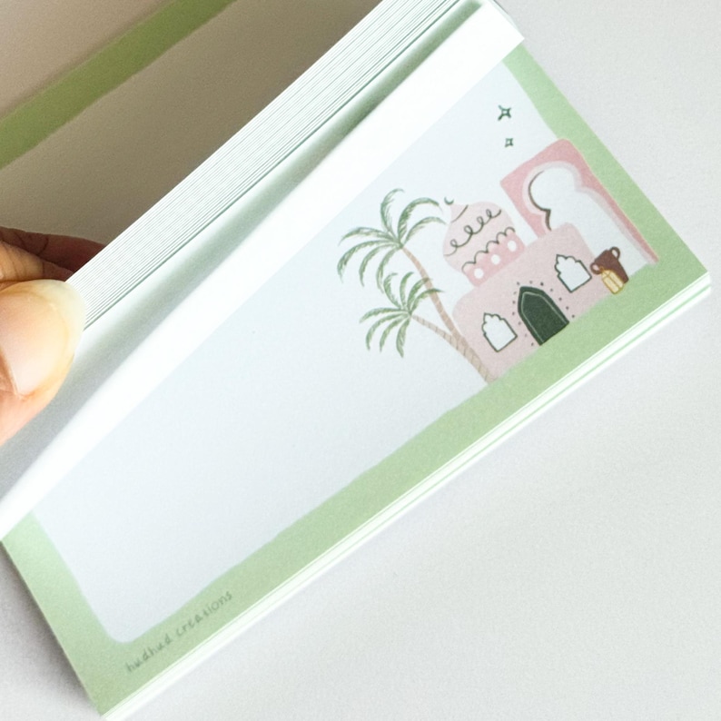 May include: A notepad with a light green border and a decorative illustration of a pink building, palm trees, and a small animal. The notepad is open, revealing blank pages. The text "hudhud creations" is printed at the bottom.