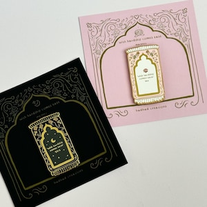 May include: Gold enamel pin with a black background featuring a prayer rug with the text "with hardship comes ease" in a gold font. The pin is on a black card with a gold border and the text "hudhud creations".
