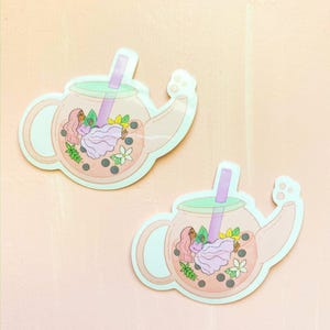 May include: Two pastel pink teapots with a green lid and a purple straw. Each teapot has a woman in a pink dress and flowers inside. The teapots are on a pink background.