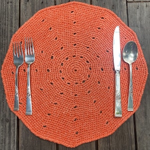 May include: A crocheted orange placemat with a circular pattern and a set of silverware, including a knife, fork, and spoon.