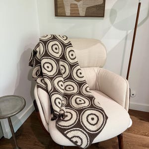 May include: A brown and white crocheted throw blanket with a circular pattern is draped over a cream-colored armchair. The chair is in a room with wood floors and a modern lamp.