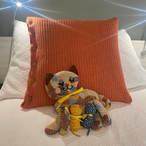 May include: A patchwork cat plush toy with two smaller patchwork kittens. The cat is sitting on a white bedspread with an orange knitted pillow behind it.