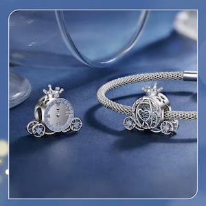 Op de afbeelding: Twee zilverkleurige bedelhangers in de vorm van een koets. De ene is een klok, de andere heeft een hartvorm. De bedels hebben een kroon en zijn versierd met kleine blauwe kristallen. Eén bedel zit aan een zilveren armband.