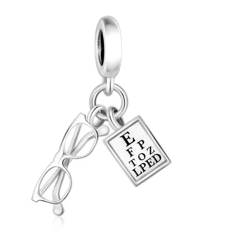 May include: Silver charm featuring a pair of eyeglasses and a miniature eye chart with the letters E, F, P, T, O, Z, L, P, E, D. The charm is designed to be attached to a bracelet or necklace, and is made of a shiny, reflective metal.
