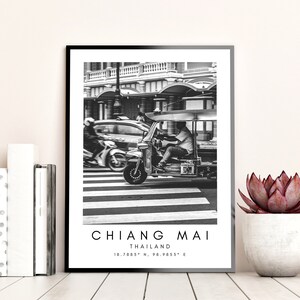 May include: Black and white photograph of a tuk-tuk driving down a street in Chiang Mai, Thailand. The tuk-tuk is a three-wheeled vehicle with a canopy and a driver in the front. The street is lined with buildings and there is a car in the background.