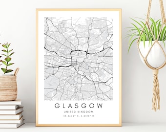 May include: A black and white map print of Glasgow, United Kingdom, with the city name and coordinates printed below the map. The print is framed in a gold frame and is displayed on a white shelf with a potted plant and a stack of books.