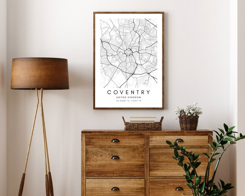 May include: A black and white map print of Coventry, United Kingdom, framed in a brown wood frame. The map shows the city's streets and landmarks. The print is hanging on a white wall above a wooden dresser.