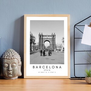 May include: Black and white photograph of the Arc de Triomf in Barcelona, Spain. The photo is framed in a light wood frame. The text "BARCELONA SPAIN 41.3851&deg; N, 2.1754&deg; E" is printed below the image.