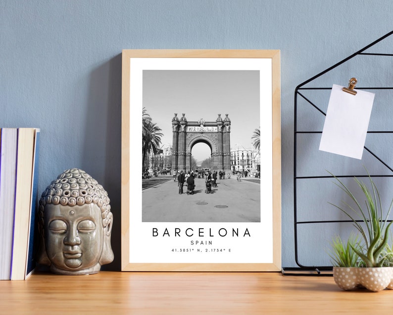 May include: Black and white photograph of the Arc de Triomf in Barcelona, Spain. The photo is framed in a light wood frame. The text "BARCELONA SPAIN 41.3851&deg; N, 2.1754&deg; E" is printed below the image.