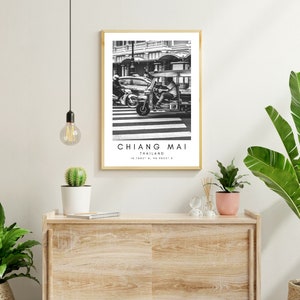 May include: Black and white photograph of a street scene in Chiang Mai, Thailand. A three-wheeled motorcycle taxi drives past a car on a crosswalk. The image is framed in a simple wooden frame.