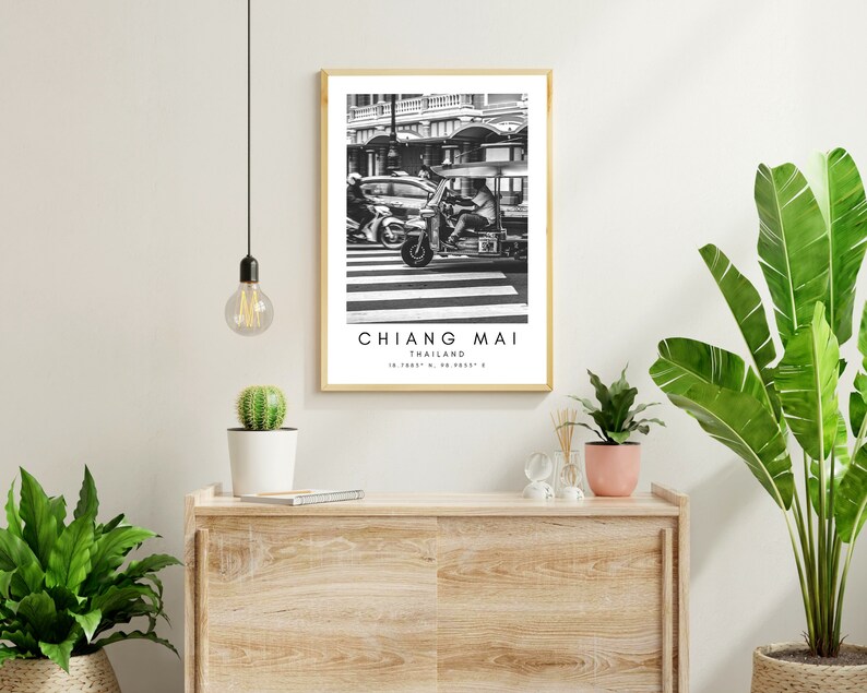 May include: Black and white photograph of a street scene in Chiang Mai, Thailand. A three-wheeled motorcycle taxi drives past a car on a crosswalk. The image is framed in a simple wooden frame.
