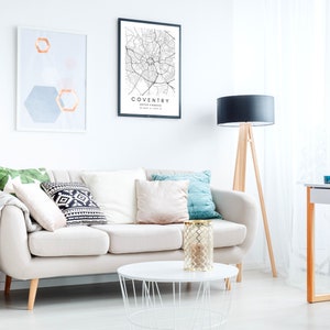 May include: A white sofa with pillows and a throw blanket in a living room with a black and white map print of Coventry, United Kingdom, a geometric print, and a black lamp with a wooden tripod stand.
