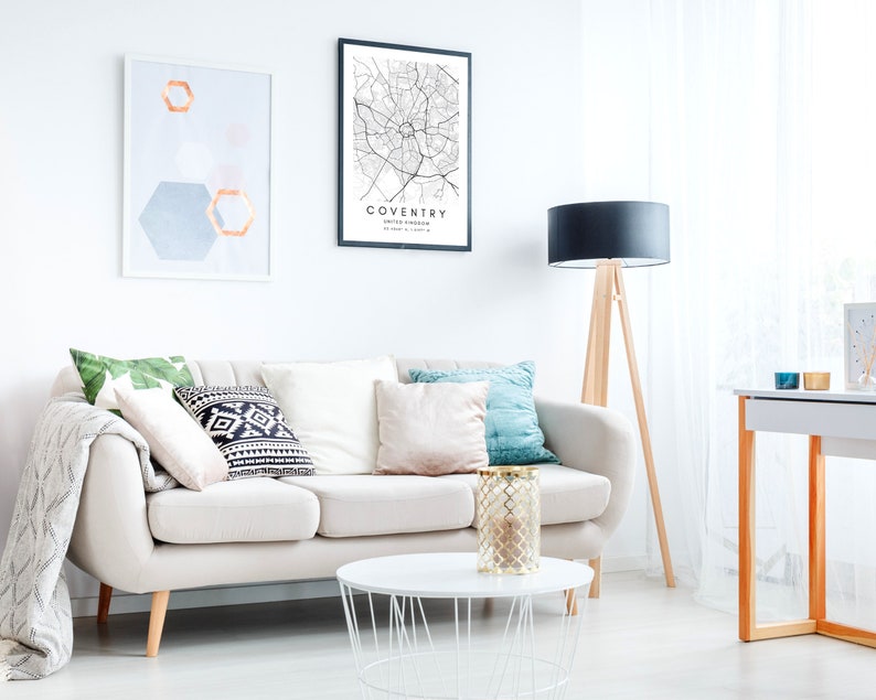 May include: A white sofa with pillows and a throw blanket in a living room with a black and white map print of Coventry, United Kingdom, a geometric print, and a black lamp with a wooden tripod stand.