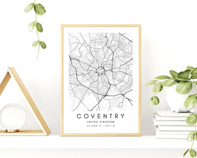 May include: A black and white map print of Coventry, United Kingdom, with the city name and coordinates printed below the map. The print is framed in a light wood frame and is displayed on a white shelf with a wooden triangle and a potted plant.