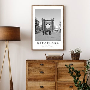 May include: Black and white photograph of the Arc de Triomf in Barcelona, Spain. The photo is framed in a simple brown frame. The text "BARCELONA SPAIN 41.3887&deg; N, 2.1754&deg; E" is printed below the image.