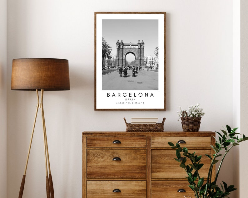 May include: Black and white photograph of the Arc de Triomf in Barcelona, Spain. The photo is framed in a simple brown frame. The text "BARCELONA SPAIN 41.3887&deg; N, 2.1754&deg; E" is printed below the image.