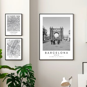 May include: Three black and white framed prints of city maps and a landmark. The top print is a map of London, the middle print is a map of New York City, and the bottom print is a photo of the Arc de Triomf in Barcelona, Spain. The prints are hanging on a white wall.