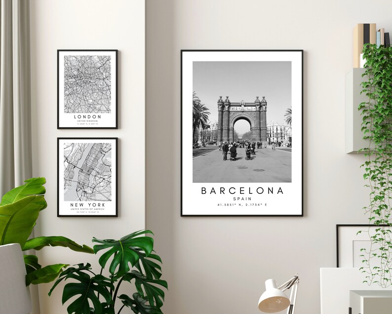 May include: Three black and white framed prints of city maps and a landmark. The top print is a map of London, the middle print is a map of New York City, and the bottom print is a photo of the Arc de Triomf in Barcelona, Spain. The prints are hanging on a white wall.