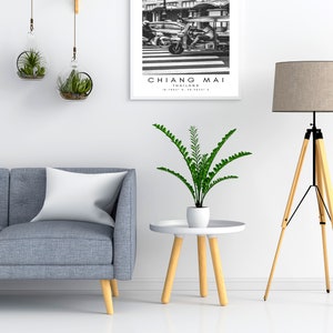 May include: A living room interior with a gray sofa, a white pillow, a potted plant, a small round wooden table, a tripod floor lamp with a beige lampshade, and a framed black and white photograph of a street scene in Chiang Mai, Thailand.